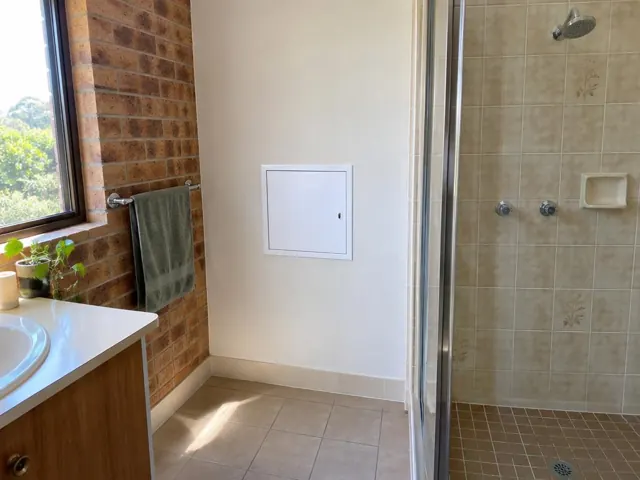 Bathroom wall with a small access panel beside a tiled shower and brick window wall in Woodberry